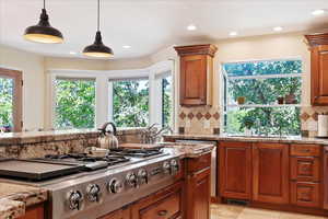 Kitchen with stainless steel gas cooktop, light stone countertops, hanging light fixtures, backsplash, and brown cabinetry
