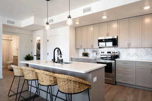 Kitchen featuring appliances with stainless steel finishes, stacked washing machine and dryer, gray cabinetry, light countertops, and an island with sink
