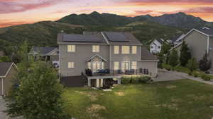 Back of property at dusk featuring solar panels, a lawn, a deck with mountain view, a chimney, and roof with shingles