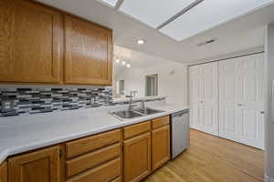 Kitchen featuring stainless steel dishwasher, decorative backsplash, brown cabinetry, light wood finished floors, and light countertops