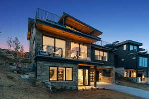 Back of house at dusk featuring stone siding, a balcony, and a patio area