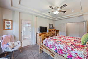 Tiled bedroom with a tray ceiling, carpet floors, ensuite bathroom, and ceiling fan