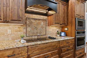 Kitchen featuring appliances with stainless steel finishes, exhaust hood, tasteful backsplash, light stone countertops, and brown cabinets