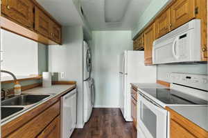 Kitchen featuring white appliances, stacked washing machine and dryer, dark wood-style flooring, brown cabinets, and light countertops