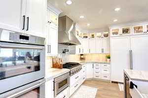 Kitchen with wall chimney exhaust hood, built in appliances, light wood-style flooring, white cabinets, and tasteful backsplash