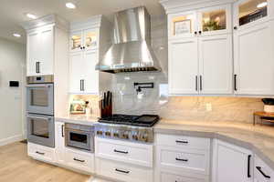 Kitchen with stainless steel appliances, wall chimney range hood, decorative backsplash, light wood-type flooring, and white cabinets