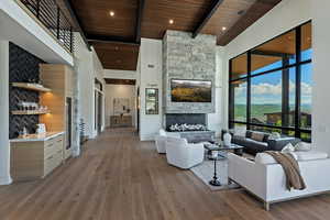 Living room featuring a towering ceiling, a stone fireplace, a wood ceiling with exposed beams, light wood finished floors, and bar area
