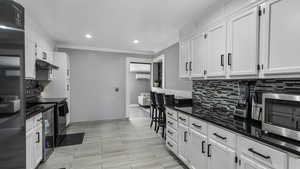 Kitchen featuring stainless steel microwave, decorative backsplash, under cabinet range hood, white cabinets, and ornamental molding
