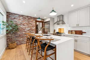 Kitchen with wall chimney exhaust hood, built in fridge, brick wall, light wood finished floors, and a center island with sink