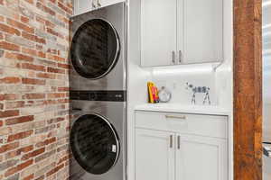 Washroom featuring stacked washing machine and dryer, brick wall, and cabinet space