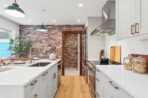 Kitchen with wall chimney exhaust hood, appliances with stainless steel finishes, brick wall, light countertops, and light wood-style flooring