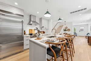 Kitchen featuring built in fridge, wall chimney exhaust hood, light wood-style floors, backsplash, and a kitchen bar