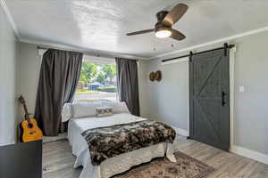 Bedroom with ornamental molding, a textured ceiling, a barn door, light wood-style flooring, and ceiling fan