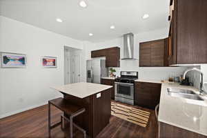 Kitchen featuring stainless steel appliances, wall chimney exhaust hood, a center island, a breakfast bar, and dark brown cabinets
