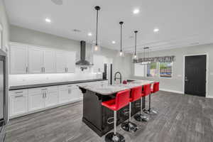 Kitchen with wall chimney range hood, backsplash, light wood-type flooring, white cabinetry, and recessed lighting