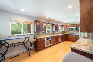 Kitchen featuring appliances with stainless steel finishes, tasteful backsplash, light stone countertops, light wood-type flooring, and glass insert cabinets