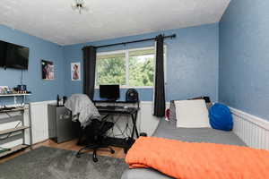Bedroom featuring wood finished floors, a textured ceiling, an office area, a textured wall, and wainscoting
