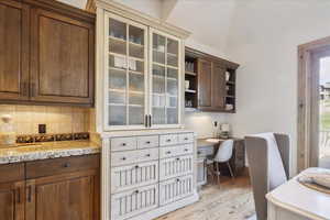 Kitchen featuring built in study area, light wood-style flooring, decorative backsplash, light stone counters, and glass insert cabinets