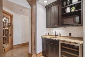 Indoor wet bar featuring beverage cooler, dark brown cabinetry, light stone countertops, open shelves, and black microwave