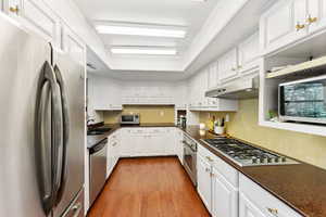 Kitchen featuring stainless steel appliances, under cabinet range hood, dark countertops, light wood finished floors, and white cabinetry