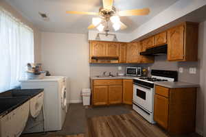 Kitchen with white gas stove, stainless steel microwave, under cabinet range hood, washer / clothes dryer, and dark wood-type flooring