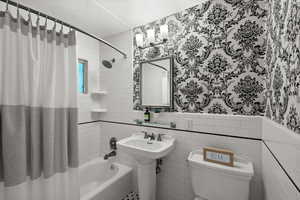 Bathroom with wallpapered walls, shower / bath combo, wainscoting, and tile walls