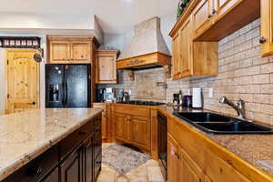 Kitchen featuring black appliances, custom range hood, decorative backsplash, and light stone countertops