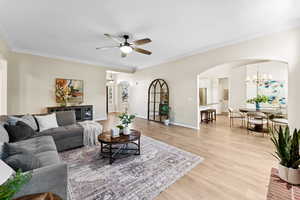 Living room with arched walkways, a ceiling fan, light wood-style flooring, a chandelier, and crown molding