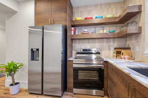 Kitchen with stainless steel appliances, open shelves, decorative backsplash, light stone counters, and light wood-style flooring