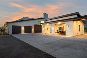Back of house at dusk featuring concrete driveway, an attached garage, stucco siding, and a chimney