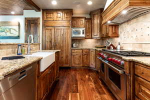 Kitchen featuring stainless steel appliances, custom exhaust hood, decorative backsplash, dark wood-style flooring, and brown cabinetry