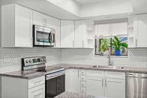 Kitchen featuring appliances with stainless steel finishes, open shelves, white cabinets, tasteful backsplash, and light stone counters