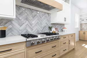 Kitchen with stainless steel gas stovetop, tasteful backsplash, glass fronted cabinets, light wood-type flooring, and light stone countertops