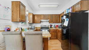 Kitchen with freestanding refrigerator, under cabinet range hood, stainless steel electric range, a peninsula, and light wood finished floors