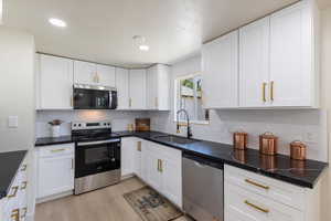Kitchen featuring stainless steel appliances, tasteful backsplash, white cabinetry, light wood-style flooring, and dark countertops