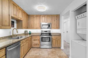 Kitchen featuring stainless steel appliances, stacked washer and clothes dryer, stone countertops, a textured ceiling, and brown cabinetry