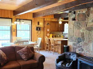 Carpeted living room with a wood stove, wood walls, a wooden ceiling with exposed beams, and ceiling fan