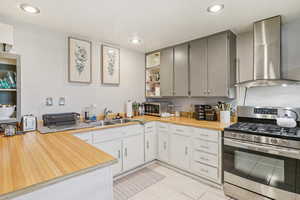 MIL - Kitchen with stainless steel gas stove, wall chimney range hood, open shelves, gray cabinets, and light tile patterned floors