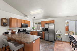 Kitchen with stainless steel appliances, a peninsula, lofted ceiling, brown cabinets, and dark countertops