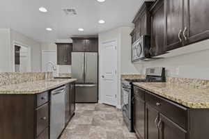 Kitchen featuring appliances with stainless steel finishes, dark brown cabinetry, light stone counters, recessed lighting, and an island with sink