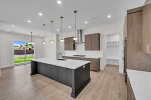 Kitchen featuring wall chimney range hood, light wood-type flooring, backsplash, a large island with sink, and recessed lighting