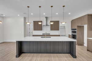 Kitchen with wall chimney range hood, double oven, light wood finished floors, decorative backsplash, and a large island with sink