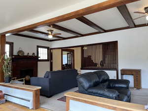 Living room featuring a ceiling fan, beam ceiling, a baseboard radiator, a brick fireplace, and light colored carpet