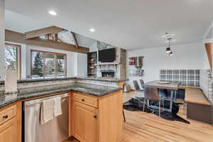 Kitchen featuring dishwasher, light wood finished floors, a stone fireplace, dark stone countertops, and recessed lighting