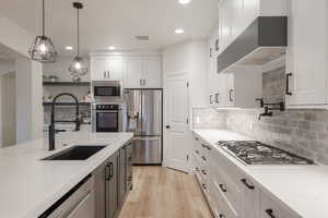 Kitchen with appliances with stainless steel finishes, wall chimney exhaust hood, white cabinetry, light wood finished floors, and decorative light fixtures