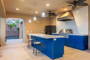 Kitchen with ventilation hood, black fridge with ice dispenser, blue cabinets, concrete flooring, and recessed lighting
