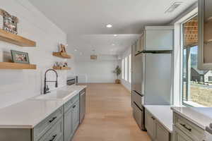 Basement kitchen with open shelves, freestanding refrigerator, stainless steel dishwasher, light wood finished floors, and gray cabinetry