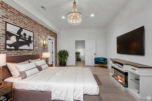 Bedroom featuring light wood-type flooring, brick wall, a chandelier, a glass covered fireplace, and recessed lighting