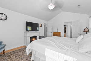 Bedroom featuring wood finished floors, a warm lit fireplace, lofted ceiling, and a ceiling fan