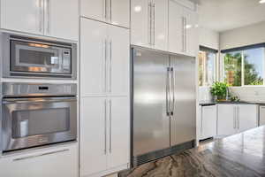 Kitchen with built in appliances, white cabinetry, backsplash, and dark stone countertops